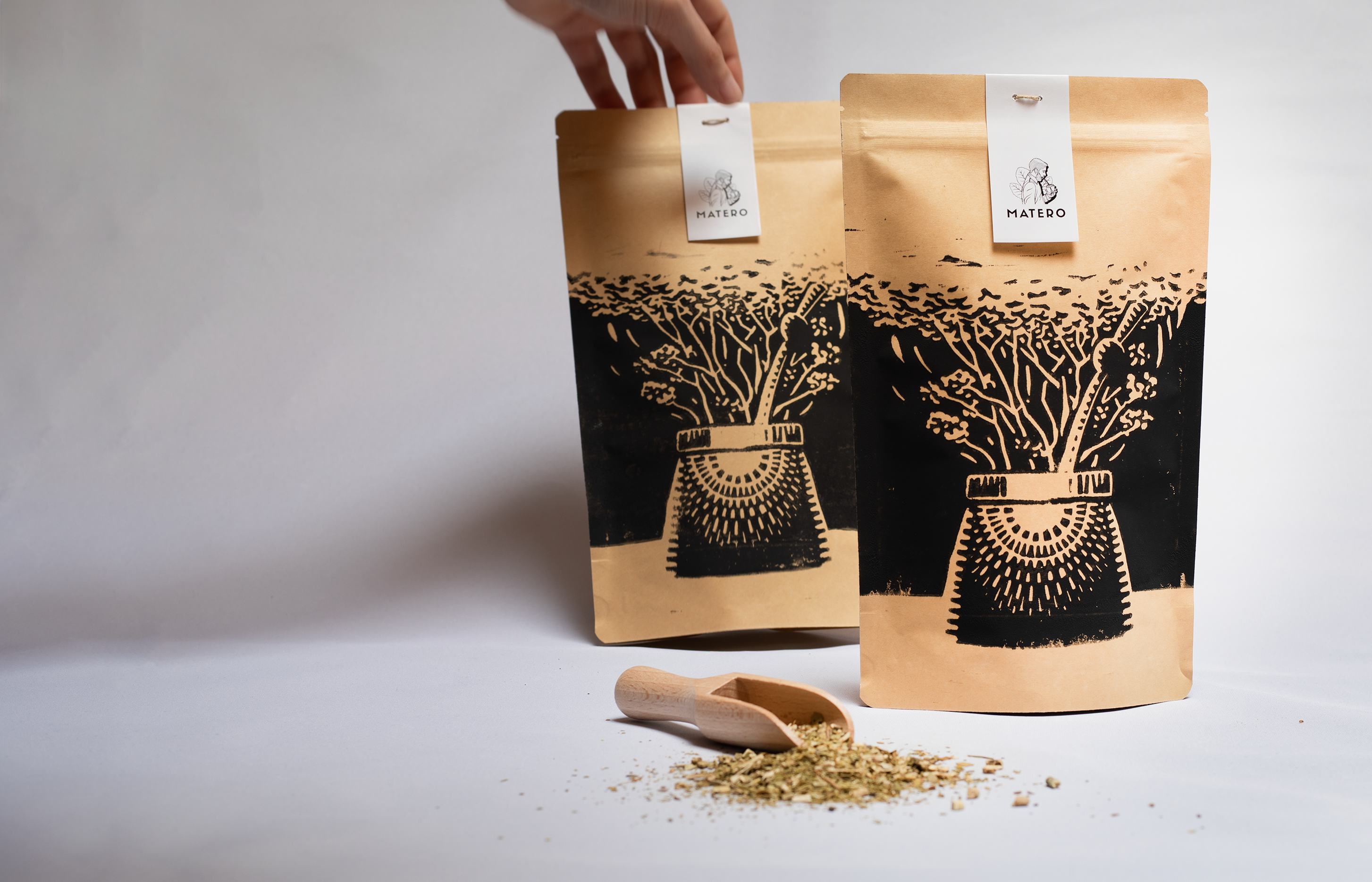 Two kraft paper pouches of Matero Yerba Mate stand on a white surface with a small wooden scoop and loose tea leaves in front. A hand is visible holding the top of the left pouch.